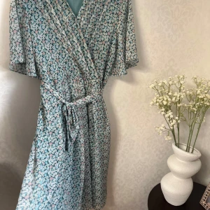 Blommig omlottklänning från Romantic Line - Supersöt blommig omlottklänning från Romantic Line i ljusa toner av blått, vitt och rosa. Klänningen har korta ärmar, knytband i midjan och v-ringning. Perfekt för dig som gillar romantiska och luftiga plagg med sommarkänsla.