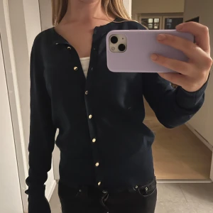 Mörkblå kofta från Lindex - Mörkblå kofta från Holly & Whyte med guldknappar framtill. Klassisk rund halsringning och ribbade muddar vid ärmslut och nederkant. Perfekt att slänga över en topp för en chill look.