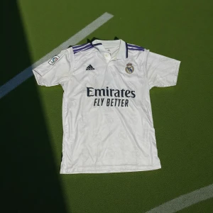 Real Madrid Benzema #9 matchtröja - Säljer en vit Real Madrid fotbollströja från Adidas med Benzema och nummer 9 på ryggen. Tröjan har korta ärmar, lila detaljer på axlarna och broderat klubbmärke på bröstet. Materialet är lätt och andas, perfekt för träning eller match. La Liga-märke på ärmen.