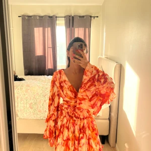 Röd och orange volangklänning med v-ringning - Säljer en färgstark kortklänning med v-ringad hals och stora volangärmar. Klänningen har ett unikt mönster i rött och orange, omlottliv och markerad midja. Materialet känns lätt och luftigt, perfekt för dig som gillar att sticka ut med din stil.