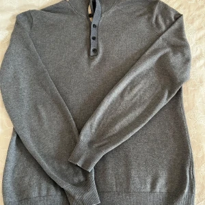 Grå stickad tröja Massimo Dutti - Snygg grå stickad tröja från Massimo Dutti i bomull och kashmir. Tröjan har lång ärm, ribbade muddar och en stilren knappslå vid halsen. Perfekt för dig som gillar en clean och klassisk look med en modern touch.