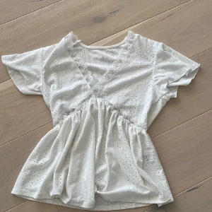 Vit spetsblus med v-ringning - Supersöt vit blus med broderade hålmönster och v-ringning med spetskant. Modellen har korta ärmar och en luftig, lätt utställd passform under bysten. Perfekt för vår och sommar när du vill ha en romantisk och fräsch look.