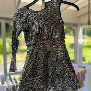 Glittrig silverklänning med volang - Snygg kort glittrig klänning i silver med asymmetrisk design och volangdetalj över bröstet. Ena ärmen är lång medan den andra sidan är ärmlös, vilket ger en cool och unik look. Perfekt för dig som vill sticka ut på festen!