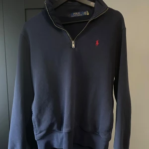 Mörkblå tröja med dragkedja från Polo Ralph Lauren - Säljer en mörkblå långärmad tröja från Polo Ralph Lauren med halv dragkedja och röd broderad logga på bröstet. Klassisk och stilren modell med mjukt material och ribbade muddar.