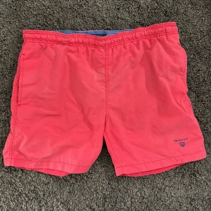 Röda badshorts från Gant - Snygga röda badshorts från Gant med blå logotyp broderad på vänster ben. Shortsen har elastisk midja och bakficka med lock. Perfekta för sommaren och strandhäng.