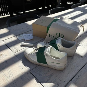 Valentino Garavani sneakers med grön detalj - Säljer ett par vita Valentino Garavani sneakers med bred grön rem över mitten, skorna är i befintligt skick och det förekommer defekter runt främre delen av skorna. Skorna har rund tå och platt sula. Perfekta inför sommaren. Även dustbag, extra skosnören, låda och original kvitto fås med vid köp. Kom med förslag! Möjligtvis öppen för byten