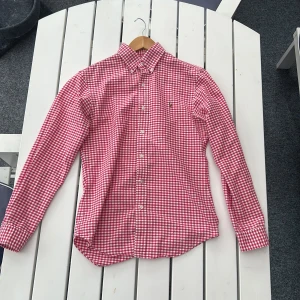 Rödvit rutig skjorta från Ralph Lauren - Säljer en röd och vit rutig skjorta från Ralph Lauren i slim fit-modell. Skjortan har klassisk krage, knäppning framtill och broderad logga på bröstet. Perfekt för dig som gillar stilrena och tidlösa plagg.
