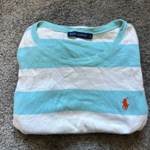 Randig tröja från Ralph Lauren - Säljer en fräsch tröja från Ralph Lauren i ljusblått och vitt med breda ränder. Tröjan har rund halsringning och klassisk passform. Den ikoniska loggan i orange finns broderad på bröstet.