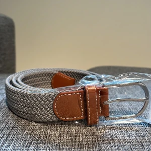 Grått flätat bälte  - Snyggt grått flätat bälte med bruna skinn-detaljer och silverfärgat spänne. Bältet har ett stilrent mönster och passar perfekt till jeans eller chinos. Klassisk design med tydliga sömmar på skinnpartierna.