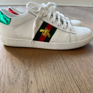 Gucci Ace vita sneakers med bi-detalj - Säljer ett par vita Gucci Ace sneakers med klassiska gröna och röda ränder samt broderad guldfärgad bi på sidan. Baksidan har metallicröd och metallicgrön häl med Gucci-logga. Skorna har snörning och platt sula.