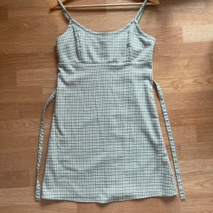 Grönrutigt klänning med smala axelband  - Säljer en söt grön och vit rutig klänning från BM. Klänningen har smala axelband, knytband i midjan och dragkedja på ryggen. Perfekt för sommaren eller picknick dag👒, enkel att styla med sneakers eller sandaler. Endast använt enstaka gånger. 💖