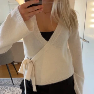 Vit omlott kofta med knytband - Säljer en supermjuk och fluffig vit omlott kofta med knytband i sidan. Koftan har lång ärm, v-ringning och är croppad i modellen. Perfekt att styla över ett linne eller topp.