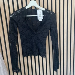 Svart spetsblus från Gina Tricot - Säljer en svart långärmad spetsblus från Gina Tricot i storlek XS. Blusen har v-ringning och är helt i genombruten spets med fina blommönster. Perfekt för dig som vill ha en elegant och trendig topp.