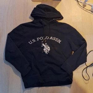 Svart hoodie från U.S. Polo Assn. - Svart hoodie från U.S. Polo Assn. med stor vit logga och polospelare framtill. Tröjan har huva med snören och långa ärmar. Klassisk och sportig stil, perfekt för en avslappnad look. Vid snabbare handel så kan priset gå ner.☺️