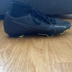 Svarta Nike Mercurial fotbollsskor - Säljer ett par svarta Nike Mercurial fotbollsskor med snörning och gula detaljer på sulan. Skorna har en högre strumpliknande krage och är designade för att ge bra grepp på planen. Perfekta för dig som vill ha stilrena och snabba skor till matchen.