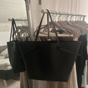 Svart axelväska i syntet - Säljer en stilren svart axelväska i syntetmaterial. Väskan har en enkel och modern design med långa handtag som gör den lätt att bära över axeln. Perfekt för att få plats med allt du behöver till vardags. Rymmer mycket och passar till många olika outfits.