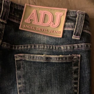 Blå jeans från Angel Devil Jeans - Säljer ett par blå jeans från Angel Devil Jeans med  ljusa partier framtill. Klassisk femficksmodell med lila sömmar och knapp med logga. Perfekta för dig som gillar Y2k/grunge/emo/ 2000’s och avslappnad stil. Jag fick ärva dessa av min mamma som hade dem under 2000 talet. Jätte fina men använder aldrig dem så bestämt mig för att sälja vidare❤️ Dem är även lågmidjade och passar (XS,S (37,36,35,34) 