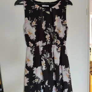Blommig klänning - Säljer en svart klänning med stora vita och ljusrosa blommor samt gröna blad. Klänningen är ärmlös och har en rund halsringning. Midjan är markerad och kjolen har ett lätt fall. Perfekt för sommarens alla tillfällen! Liten i storlek 