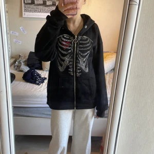Svart hoodie med strass-skelett från Romwe - 50kr Säljer en svart hoodie från Romwe med dragkedja och huva. Framsidan har ett detaljerat skelettmönster i strass och ett rött hjärta. Hoodien är i mjuk bomullsblandning och har fickor framtill. Perfekt för dig som gillar unika detaljer!