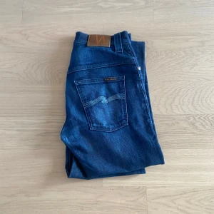 Mörkblå jeans från Nudie Jeans - Säljer ett par mörkblå jeans från Nudie Jeans med klassisk femficksdesign och raka ben. Jeansen har en snygg tvätt och detaljerade sömmar på bakfickorna samt läderpatch i midjan.obs! Dom är lite förkortade.