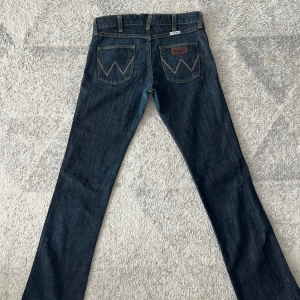 Mörkblå jeansbyxor lowaist  från Wrangler - Säljer ett par klassiska mörkblå jeans från Wrangler med raka/ bootcut ben och snygga kontrastsömmar. Byxorna har fem fickor, dragkedja och knapp framtill samt tydliga W-detaljer på bakfickorna. Den 
