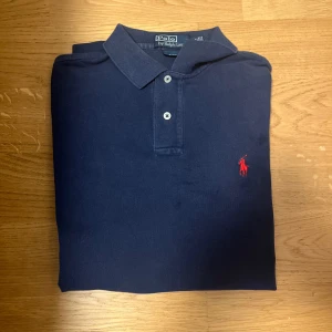 Mörkblå pikétröja från Polo Ralph Lauren - Säljer en klassisk mörkblå pikétröja från Polo Ralph Lauren med röd broderad logga på bröstet. Tröjan har krage, två knappar och är perfekt för en stilren look.