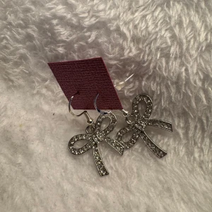 Glittriga rosettörhängen i silver - Säljer ett par stora örhängen i form av rosetter, täckta med glittrande stenar. De är i silverfärg och hängs i örat med en enkel krok. Perfekta för att ge din look lite extra bling! Krokarna är i sterling silver 