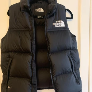 Svart dunväst från The North Face - Säljer en svart dunväst från The North Face med hög krage och dragkedja framtill. Västen har två fickor med dragkedja och logga på bröstet samt ryggen. Perfekt för lager-på-lager och håller dig varm under kyliga dagar.  OBS Tänk på att de är L i youth storlek så den sitter kanske som en XS