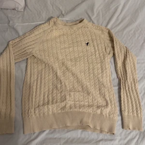 Stickad Pier One tröja - Säljer en beige stickad tröja från Pier One med broderad logga på bröstet. Tröjan har lång ärm, rund halsringning och ribbade muddar vid ärmslut och nederkant. Perfekt för lager-på-lager-stil.