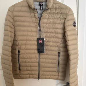 Beige pufferjacka från Colmar - Säljer en beige pufferjacka från Colmar med horisontella sömmar och två dragkedjefickor framtill. Jackan har hög krage och Colmar-logga på ärmen. Perfekt för kyliga dagar.