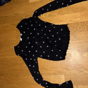 Svart kofta med broderade blommor från H&M - Säljer en svart kofta från H&M med små lila broderade blommor över hela tröjan. Koftan har långa ärmar, knappar framtill och en lätt croppad passform med vågig nederkant. Perfekt för att piffa upp din outfit!