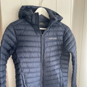 Blå lättviktsjacka från Peak Performance - Säljer en blå lättviktsdunjacka från Peak Performance med huva och dragkedja framtill. Jackan är lätt, har quiltad design och passar perfekt till vår/sommar och även höst. En Klassisk logga på bröstet och en rak passform som passar både kille och tjej. Är dom en small i dam.