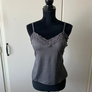 Grå linnetopp med spetskant - Säljer en stilren grå linnetopp med smala axelband och dekorativ spets längs v-ringningen. Perfekt att matcha med jeans eller kjol för en enkel och snygg look. Helt ny, har aldrig kommit till användning, axelband kan regleras!