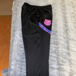 Svarta FC Barcelona träningsbyxor från Nike  - Svarta träningsbyxor med FC Barcelona-logga och Nike Swoosh på framsidan. Byxorna har elastisk midja och blå/röda mönstrade detaljer längs benen. Perfekta for fotboll eller träning.