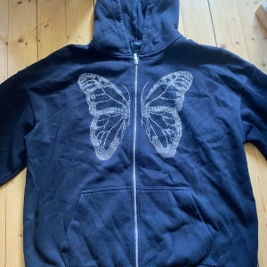 Svart hoodie med fjärilstryck - Säljer en svart hoodie med dragkedja och en stor fjäril i strass på framsidan. Tröjan har huva och fickor framtill. Perfekt för dig som gillar snygga detaljer och vill ha en bekväm stil. Ingen paljett har lossnat.