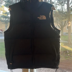 Svart dunväst från The North Face - Säljer en svart dunväst från The North Face med dragkedja framtill och hög krage. Västen har två fickor och tydlig logga på bröstet. Perfekt för lager-på-lager under kyliga dagar.