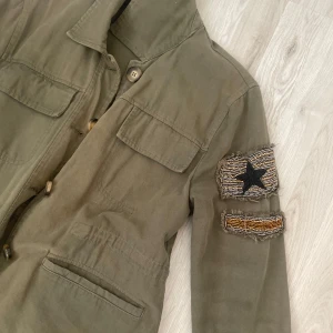 Olivgrön militärinspirerad jacka med patch - Säljer en olivgrön jacka med klassisk krage och fyra fickor framtill. Jackan har långa ärmar och en dekorativ patch med stjärna på ena ärmen. Stängs med knappar och har dragsko nedtill.