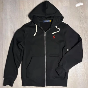 Svart zip hoodie från Polo Ralph Lauren - Säljer en svart hoodie från Polo Ralph Lauren med dragkedja och vit snörning i huvan. Tröjan har en röd broderad logga på bröstet och två fickor framtill. Perfekt för en Grisch stil. Aldrig andvänd, STORLEKEN ÄR M MEN SITTER SOM EN S.