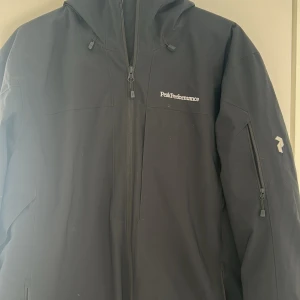 Svart jacka från Peak Performance - Säljer en svart jacka från Peak Performance med huva och dragkedja framtill. Jackan har en ficka med dragkedja på ärmen och logga på bröstet. Perfekt för dig som vill ha en stilren och funktionell jacka. Är mer som en skidjacka, men funkar till vardagligt bruk. 