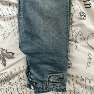 Blå jeansbyxor från Zara, storlek 34 - Säljer ett par klassiska blå jeans från Zara med raka ben och hög midja. Jeansen har knappar i silver framtill och detaljerade sömmar. Perfekta för en avslappnad och stilren look.