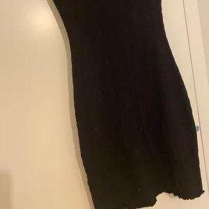 Svart kortklänning med fjärilsdetalj - Säljer en svart kortklänning med tight passform och korta ärmar. Klänningen har en liten broderad fjäril framtill vid halsen och är gjord i ett stretchigt, ribbat material. Perfekt för dig som gillar enkel men söt stil.