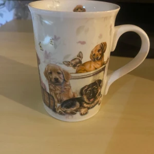 Vit mugg med hundmotiv - Säljer en söt vit porslinsmugg dekorerad med flera olika hundvalpar i färg. Muggen har blommiga detaljer, små bin och fåglar i bakgrunden. Perfekt för dig som älskar hundar och vill ha en unik mugg till kaffet eller teet.