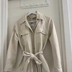 Beige overshirt från ZARA med bälte - Säljer en beige jeansjacka från ZARA med bälte i midjan och knappar framtill. Jackan har två bröstfickor med lock och långa ärmar. Perfekt för lager-på-lager och enkel att styla. knappen på högerarm har lossnat men går att sy till! 