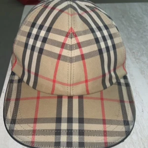 Burberry keps - Säljer en klassisk beige keps från Burberry med det ikoniska rutiga mönstret i svart, rött och vitt. Kepsen har justerbart spänne bak och är tillverkad i bomull. Perfekt för dig som gillar stilrena accessoarer med lyxig känsla.
