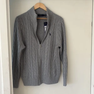 Grå stickad kofta från Polo Ralph Lauren - Säljer en grå stickad kofta från Polo Ralph Lauren med dragkedja och kabelstickat mönster. Tröjan har lång ärm, ribbade muddar och klassisk broderad logga på bröstet. Perfekt för lager-på-lager och snygg till både jeans och chinos. Jag är 189cm och väger 89kg