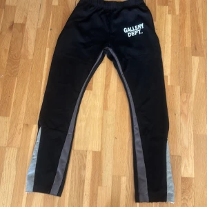 Svarta träningsbyxor från Gallery Dept. - Svarta träningsbyxor från Gallery Dept. med vitt tryck på framsidan. Byxorna har elastisk midja och gråa paneler längs benen. Perfekta för en sportig look.