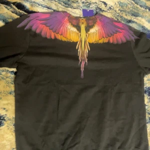 Svart tröja med vingmönster från Marcelo Burlon - Cool svart tröja från Marcelo Burlon med ikoniskt vingmönster runt halsen. Ena tröjan har färgglada vingar i lila, rosa och gult, den andra har grå och vita vingar. Perfekt statement-plagg för dig som vill sticka ut.