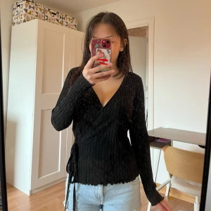 Svart omlottopp med ribbad struktur - Säljer en svart omlottopp med ribbad struktur och långa ärmar. Toppen har knyt i sidan och en djup v-ringning. Är köpt här på plick för 200 kr (har inte koll på original pris) och modellen har ofast S i tröj-storlek. Jättefin med ett linne under!!