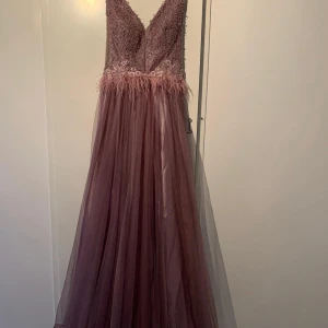 Lång lila balklänning med spets och fjädrar - Säljer en elegant långklänning i lila med v-ringning, dekorerad med spets och blommiga detaljer upptill samt fjädrar i midjan. Kjolen är i tunn, svepande tyll som ger en lyxig känsla. Perfekt för bal eller fest. har använts endast en gång. 