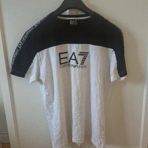 Vit och svart t-shirt från EA7 Emporio Armani - Snygg t-shirt från EA7 Emporio Armani i vit och svart med logga på bröstet och tryck längs ärmarna. Klassisk passform och korta ärmar. Perfekt för en stilren och sportig look.